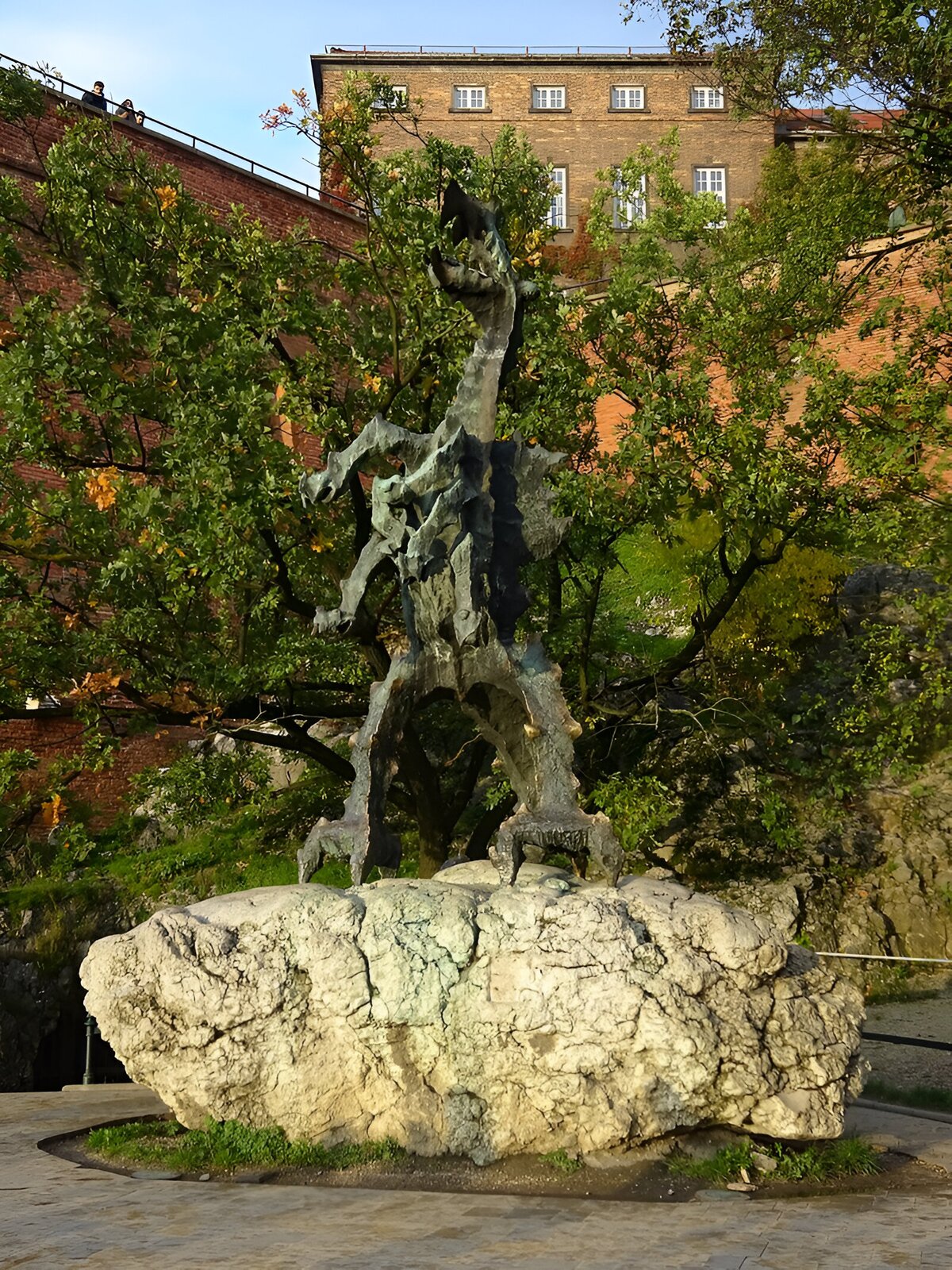 Zdjęcie przedstawia rzeźbę smoka wawelskiego na dużym szarym kamieniu. Za rzeźbą widać drzewa liściaste, mur z czerwonej cegły, miejskie zabudowania oraz bezchmurne błękitne niebo.