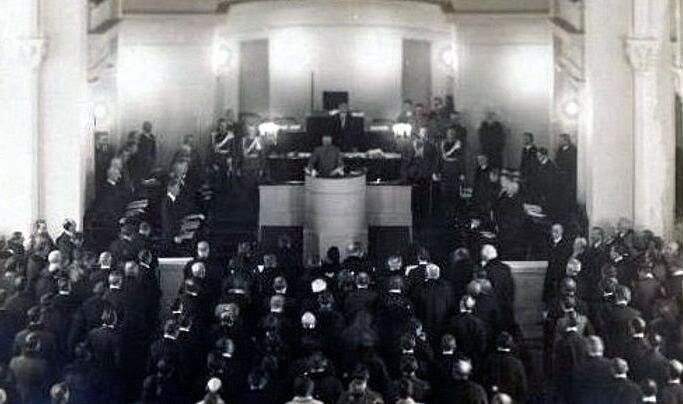 Zdjęcie przedstawia salę posiedzeń. Na środku sali stoi mównica, z której przemawia mężczyzna. Wokół niego stoją ludzie. Przed mównicą również stoją ludzie.