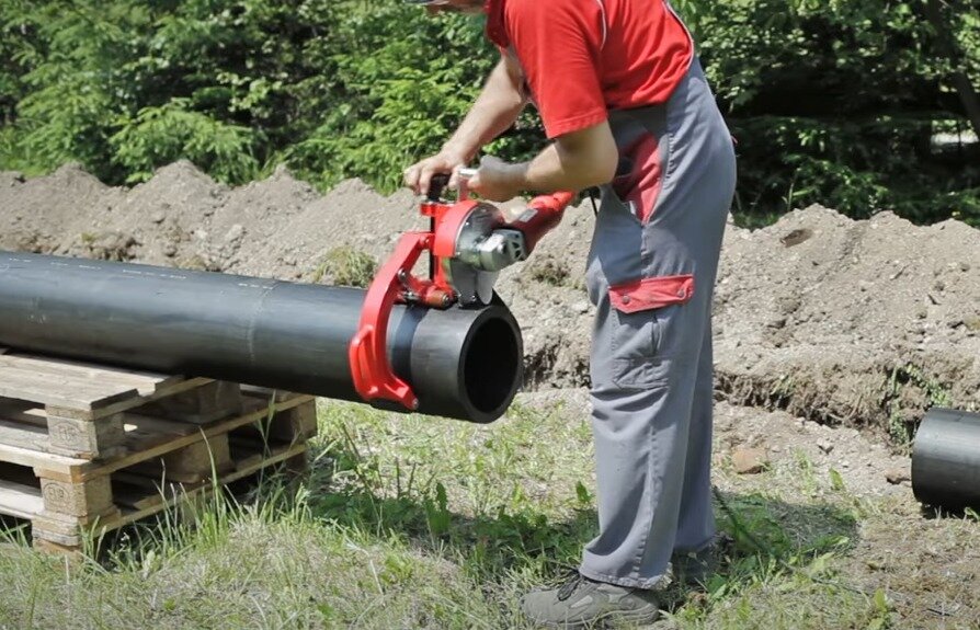 Na zdjęciu widoczny jest teren otwarty z wykopem, a na nim pracownik trzymający oburącz piłę, której ramiona oplatają rurę o dużym przekroju. Piła posiada dwa uchwyty i dwie rolki, które umożliwiają obracanie rury podczas cięcia. Poniżej ilustracji znajduje się przycisk do odtworzenia nagrania.