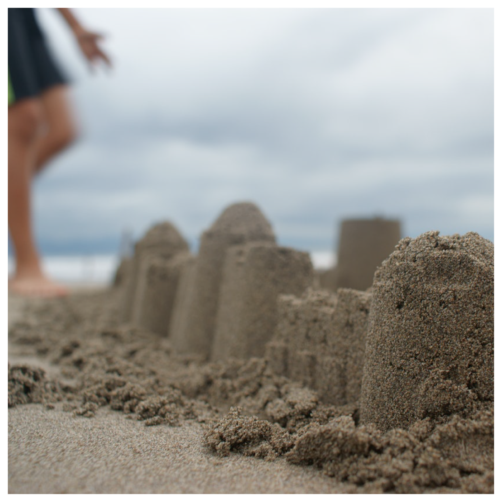 Na zdjęciu widoczne są zbudowane na plaży zamki z piasku. W tle widoczna jest osoba chodząca po plaży, morze i zachmurzone niebo. 