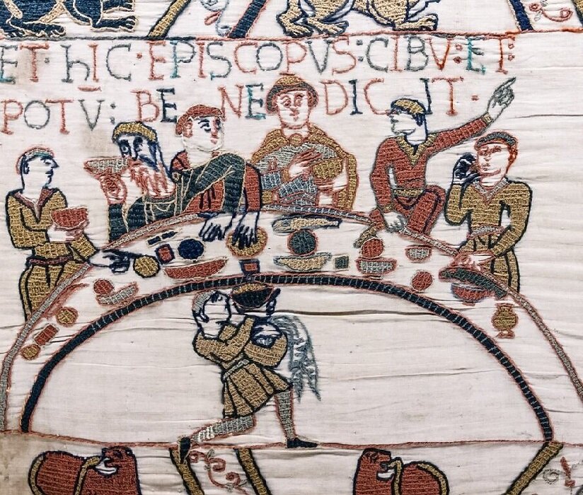 Ilustracja przedstawia mężczyzn biesiadujących przy okrągłym stole, na którym znajdują się liczne potrawy. Wokół stołu chodzą służący z naczyniami. Mężczyźni dyskutują.