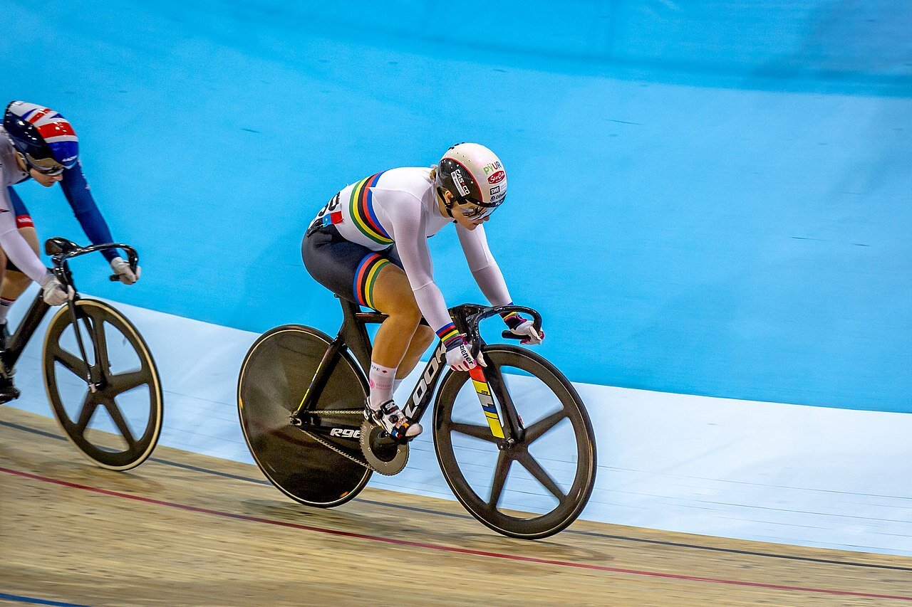Zdjęcie przedstawia specjalny tor kolarski we wnętrzu hali sportowej . Na pierwszym planie znajduje się para zawodników, którzy są ubrani w białe stroje, noszą kaski oraz okulary ochronne. 
