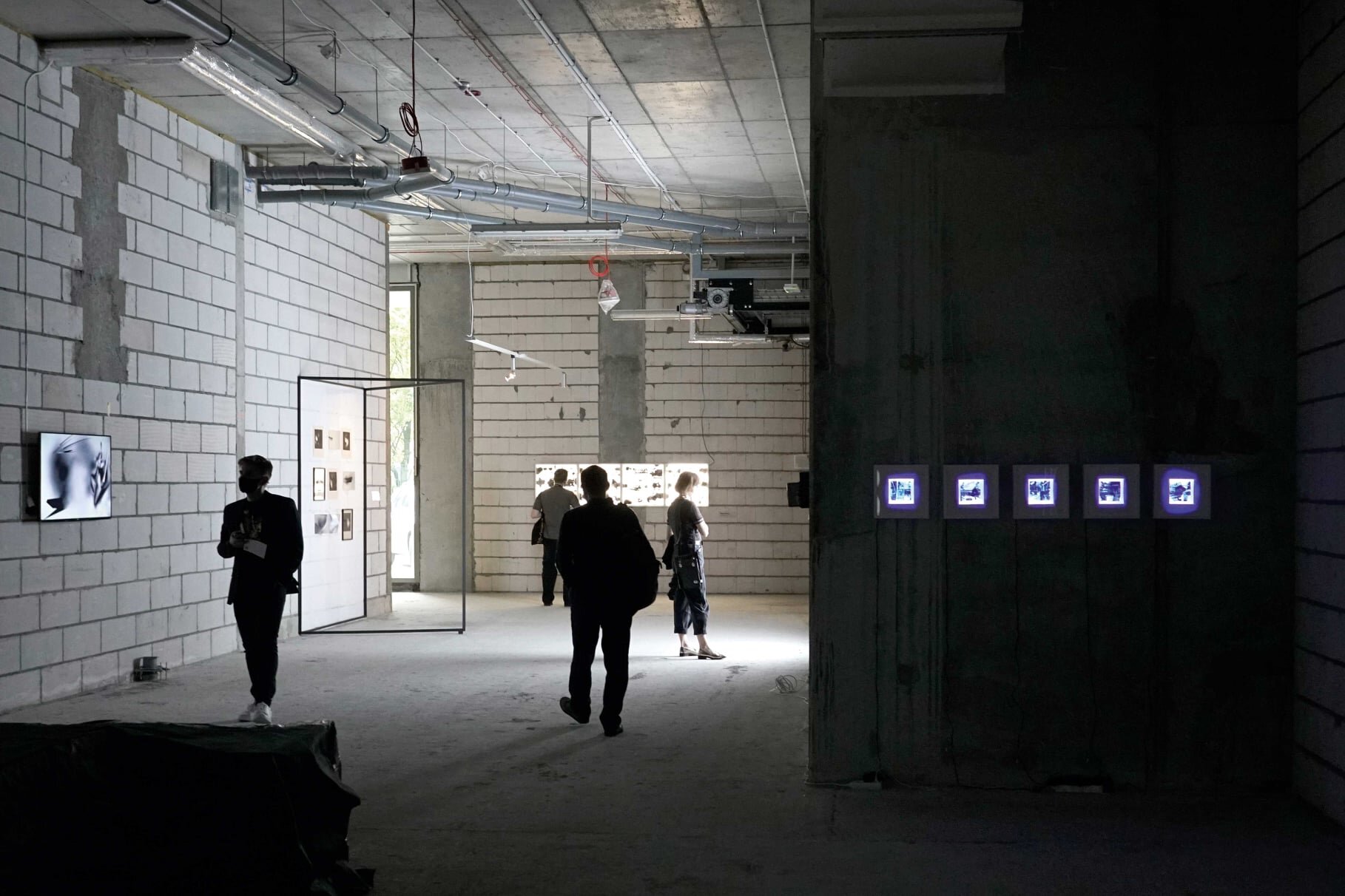 Zdjęcie jest w kolorystyce biało‑czarnej, przedstawia wysokie pomieszczenie o surowych ścianach. Pod sufitem zamocowane są rury i okablowanie. Po prawej stronie na ciemnej ścianie znajduje się rząd pięciu podświetlonych zdjęć w kształcie kwadratów. W pomieszczeniu znajdują się 4 osoby.