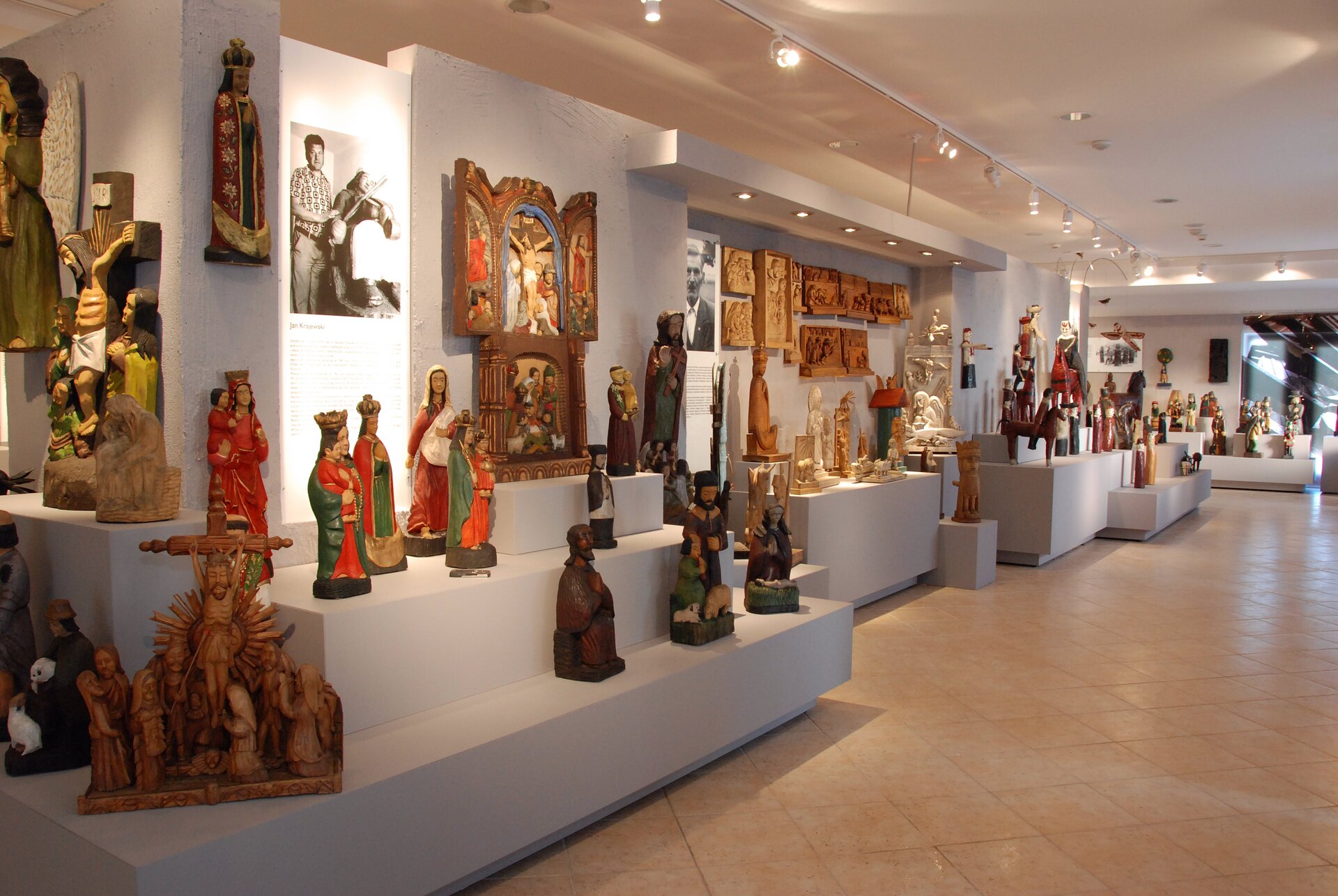 Zdjęcie przedstawia wystawę rzeźb ludowych, ustawionych na różnych poziomach przy białej ścianie. Rzeźby są drewniane, a niektóre są do tego pomalowane. Tematyka jest w głównej mierze religijna. Rzeźby przedstawiają między innymi sceny ukrzyżowania Jezusa, modlące się postaci, Maryję z małym dzieckiem na ramieniu.
