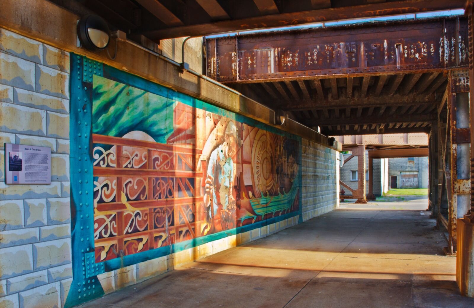 Zdjęcie przedstawia kryte przejście między blokami, którego ścianę pokryto muralem. Malowidło nie jest dobrze wyeksponowane, bo zdjęcie zostało zrobione z boku. Początek ściany został pomalowany w ułożone regularnie kamienne ciosy, za nimi dekoracyjne ornamenty ujęte w płytki, dalej obraz, składający się z różnych płaszczyzn, wśród których można wyróżnić coś na kształt starożytnej łodzi.