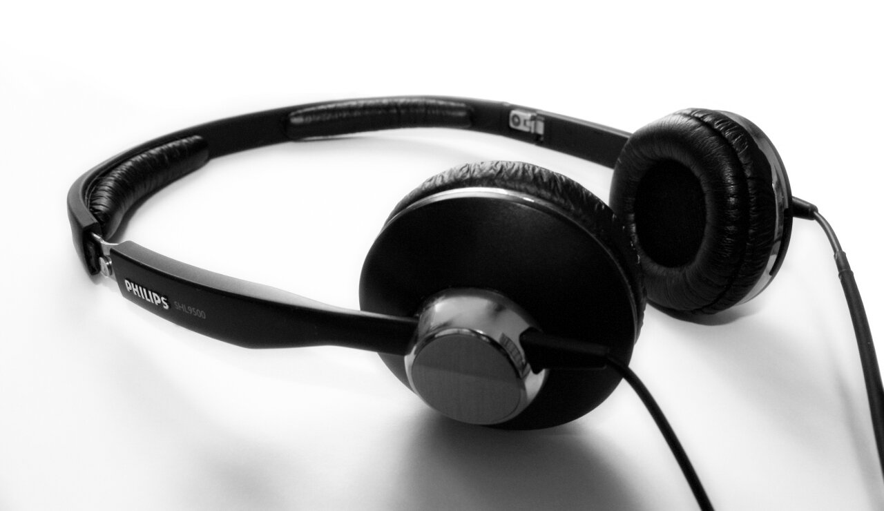 Zdjęcie przedstawia słuchawki odsłuchowe półotwarte. Widoczne są dwa elementy – nauszniki, skierowane ku sobie, z czarną częścią wewnętrzną pustą w środku oraz czarną opalizującą częścią zewnętrzną, do której przytwierdzona jest półkolista podwójna czarna obejma. Na zewnętrznej stronie lewego nausznika widoczny szary walcowaty element – obrazujący miejsce, z którego wychodzi czarny kabel, i do którego zamocowana jest obejma. Od prawego nausznika odchodzi w dół czarny kabel.