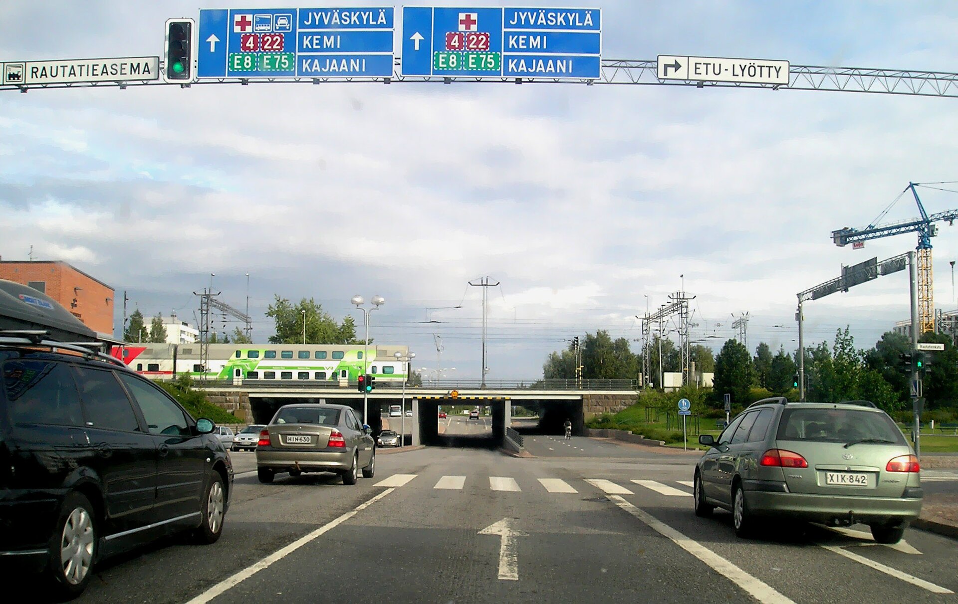 Zdjęcie przedstawia trzypasmową ulicę, na której znajduje się kilka samochodów. Nieco dalej wiadukt kolejowy z jadącym pociągiem. Nad jezdnią, na słupie znajdują się drogowskazy w języku fińskim. 