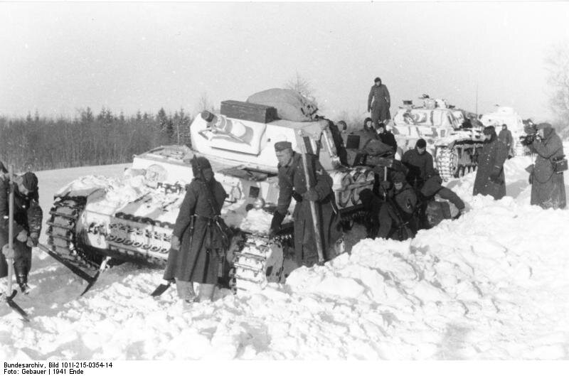 Na zdjęciu niemieccy są żołnierze ubrani w długie płaszcze. Stoją albo kucają obok trzech czołgów. Czołgi stoją jeden za drugim na zaśnieżonej drodze. Wokół nich są zaspy śniegu. W oddali widać las.