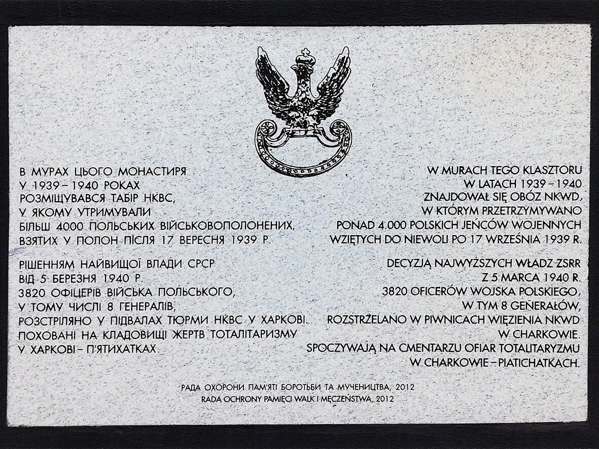 Zdjęcie przedstawia tablicę pamiątkową, na której znajduje się symbol wojskowy orła w koronie oraz napisy w języku polskim i rosyjskim. Napis polski brzmi: W murach tego klasztoru w latach 1939-1940 znajdował się obóz NKWD, w którym przetrzymywano ponad cztery tysiące polskich jeńców wojennych wziętych do niewoli po 17 września 1939 roku. Decyzją najwyższych władz ZSRR z 5 marca 1940 roku trzy tysiące dwudziestu oficerów wojska polskiego, w tym ośmiu generałów, rozstrzelano w piwnicach więzienia NKWD w Charkowie. Spoczywają na cmentarzu ofiar totalitaryzmu w Charkowie - Piatichatkach. Rada Ochrony Pamięci i Walki i Męczeństwa 2012. 