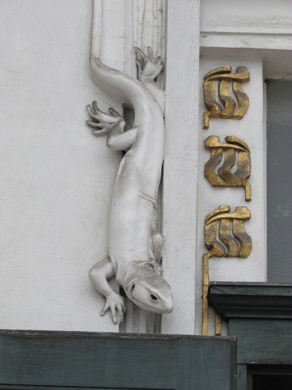 Ilustracja przedstawia detal widoczny na elewacji Pawilonu Secesji Wiedeńskiej. Detal przedstawia jaszczurkę, której autorem jest Josef Maria Olbrich. Jaszczurka jest koloru srebrnego i schodzi po ścianie. Obok jaszczurki widoczne są złote ornamenty.