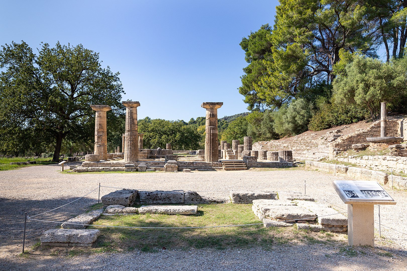 Na zdjęciu plac wysypany drobnymi kamieniami, pośrodku których starożytne zabudowania. Kamienne ruiny, kolumny. Po prawej części kamienne trybuny, dookoła drzewa.