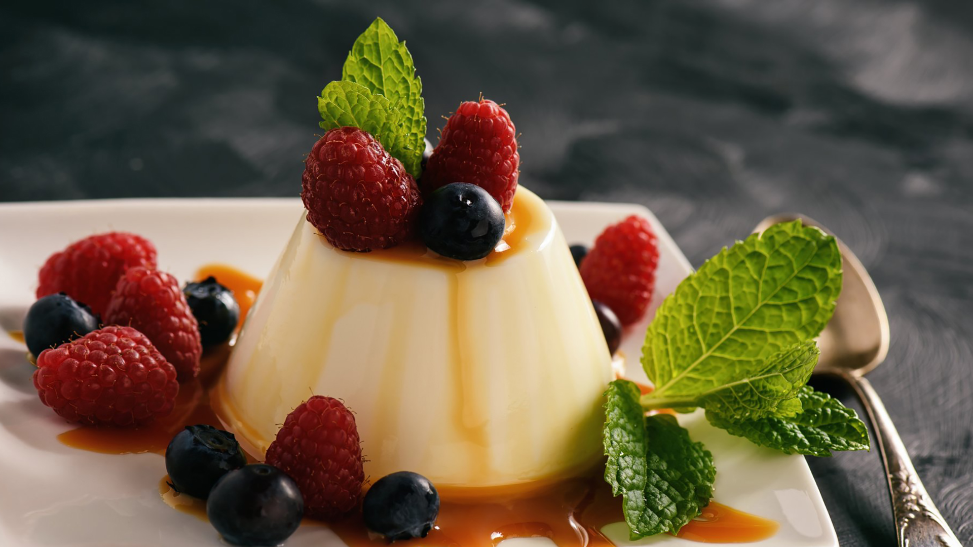 Zdjęcie przedstawia włoski deser panna cotta. Mleczny deser jest w formie babeczki. Na wierzchu deseru znajdują się maliny, jagody oraz listek mięty. Obok deseru na talerzu leżą również maliny, jagody. Deser przybrany jest listkami mięty.