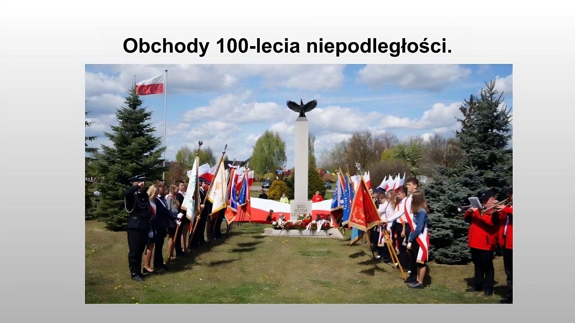 Jasnoszary slajd. Na górze slajdu nagłówek: „Obchody stulecia niepodległości”. Poniżej zdjęcie przedstawiające szary pomnik przypominający obelisk. Na szczycie czarna rzeźba ptaka z rozłożonymi skrzydłami. W dolnej części pomnika napis: „Bóg, honor, ojczyzna”. Pod pomnikiem wieńce z kwiatów. Przed pomnikiem szpaler złożony z młodzieży ubranej na galowo, z biało czerwonymi szarfami. Niektóry trzymają kolorowe sztandary. Po lewej stronie salutuje mężczyzna w granatowym, galowym mundurze. W prawym dolnym rogu kadru wyłania się część orkiestry. Dwaj mężczyźnie w czarnych spodniach i czerwonych marynarkach grają na trąbkach. Z pomnikiem stoją ludzie trzymający dużą flagę polski. Po lewej stronie za pomnikiem na maszcie powiewa flaga polski. Na około zielona trawa i drzewa. 
