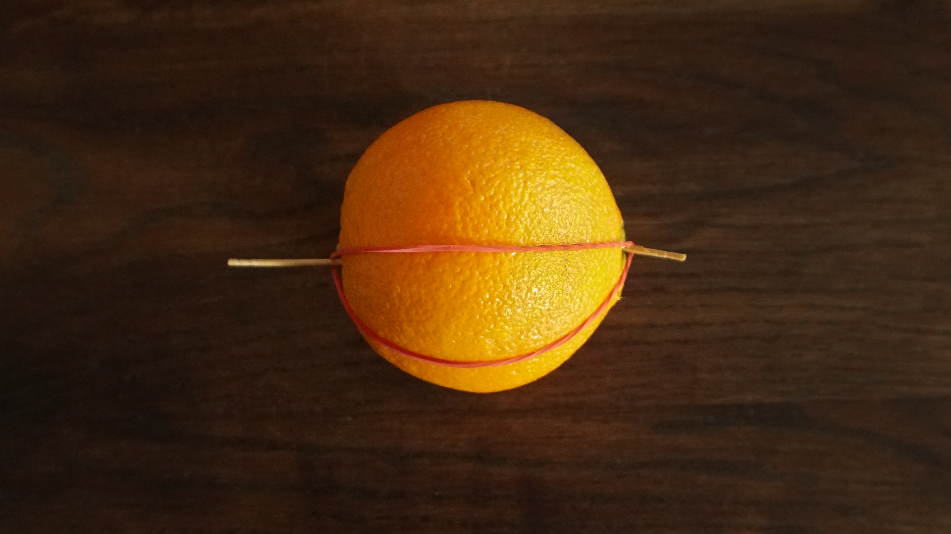 Zdjęcie przedstawia pomarańczę. Na obu biegunach wbito po jednej zapałce i założono na gumkę recepturkę. Gumka tworzy figurę w kształcie oka.