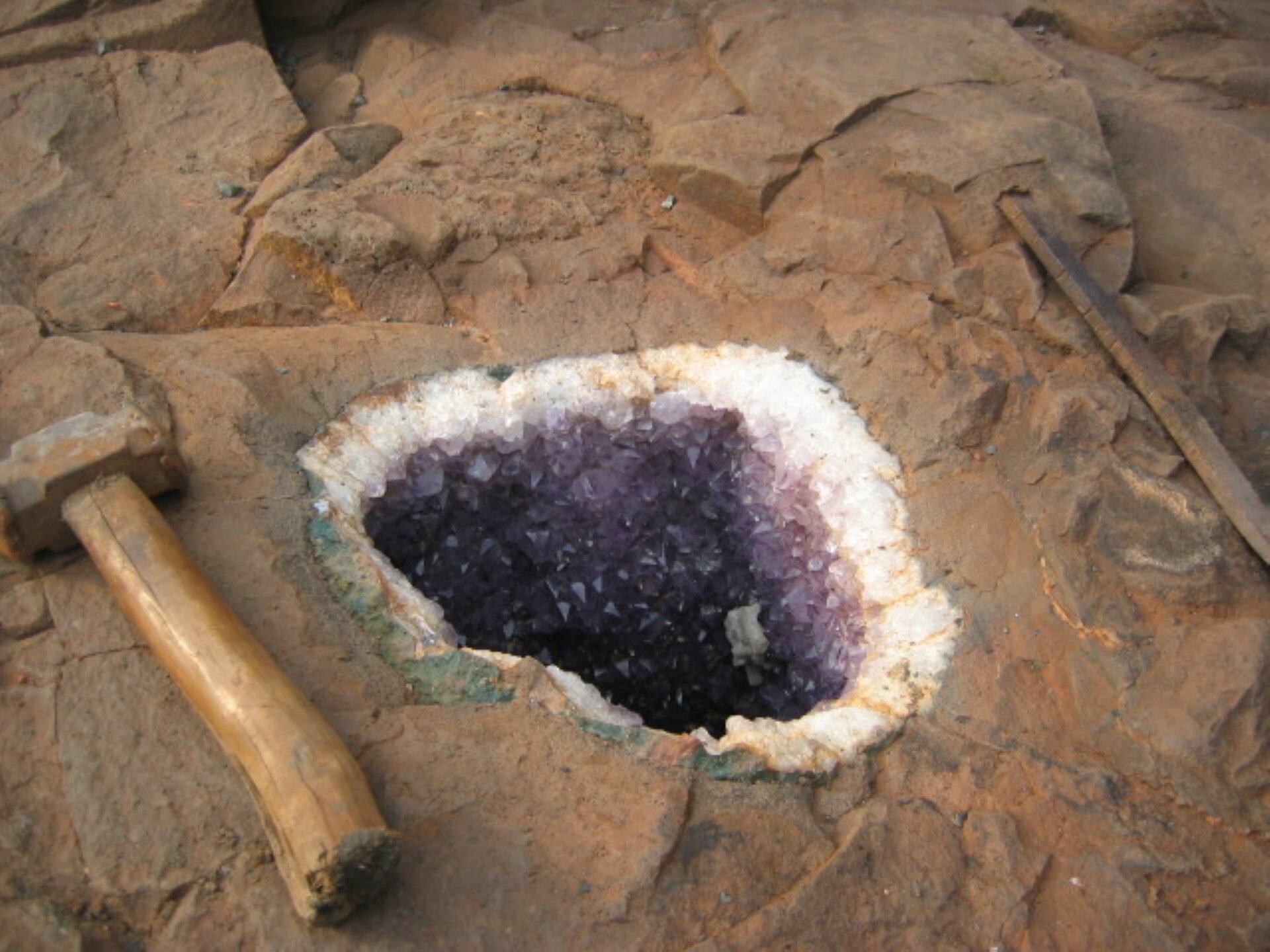 Rys. a. Zdjęcie przedstawia skałę o barwie szaro‑brązowej. Pośrodku skały wyrąbano otwór, którego brzegi są białe i błyszczące, a centrum wypełniają jasno i ciemno‑fioletowe kryształki, odbijające światło. 