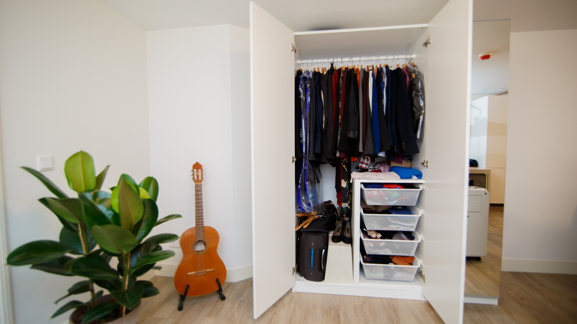 Zdjęcie przedstawia pokój, w którym widoczna jest otwarta szafa, a w niej poukładane ubrania na wieszakach. Po lewej stronie szafy stoi gitara i kwiat.