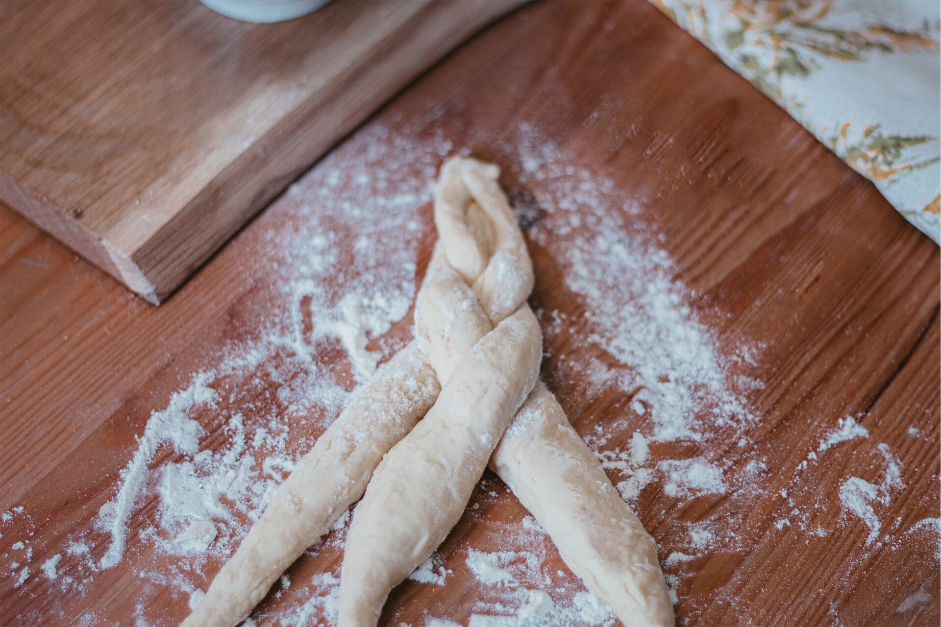 Zdjęcie przedstawia krok czwarty i piąty formowania chały tureckiej. Ujęcie wykonane jest od góry pod skosem. Widoczny jest ciemnobrązowy drewniany blat na którym w środkowej części grafiki znajdują się trzy podłużne owalne wałki ciasta oprószone białą mąką. Końce wałków z jednej strony są ze sobą złączone, a w pozostałej części długości wałki wzajemnie się oplatają na krzyż. Dookoła wałków rozsypana jest mąka. W prawej górnej części zdjęcia dostrzec można biały obrus z zielonymi i żółtymi motywami roślinnymi. W lewym górnym rogu grafiki widoczna jest jasnobrązowa drewniana deska ułożona pod skosem.