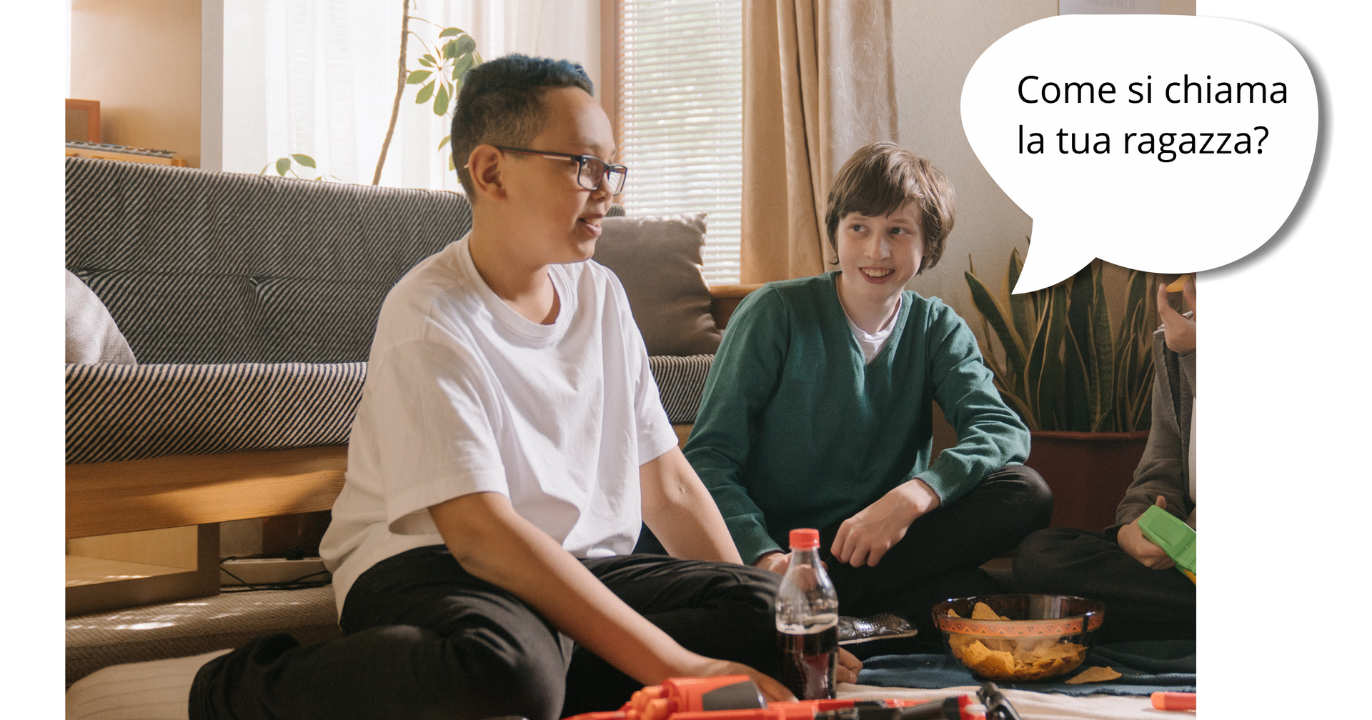 Zdjęcie przedstawia dwóch nastoletnich chłopaków siedzących na podłodze w pokoju. Chłopak po lewej nosi okulary. Chłopak po prawej zwraca się w stronę drugiego chłopca, uśmiecha się. Obok niego znajduje się dymek z pytaniem: "Come si chiama la tua ragazza?"