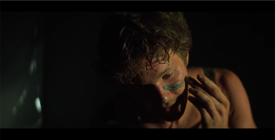 Kadr przedstawia mężczyznę, który maluje zieloną farbą poziome kreski na twarzy pod oczami. Mężczyzna ma krótkie włosy, wokół niego panuje ciemność.