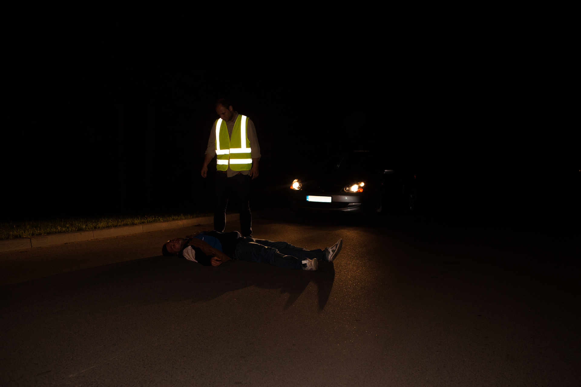 Noc. Na jezdni leży nieprzytomna osoba. Leżąca osoba bez odblaskowych dodatków. Za leżącą osobą zaparkowany samochód z włączonymi reflektorami. Reflektory skierowane na leżącą osobę. Kierowca w żółtej kamizelce odblaskowej wyróżnia się na ciemnym zdjęciu. Białe pasy na kamizelce ostrzegają innych kierowców.