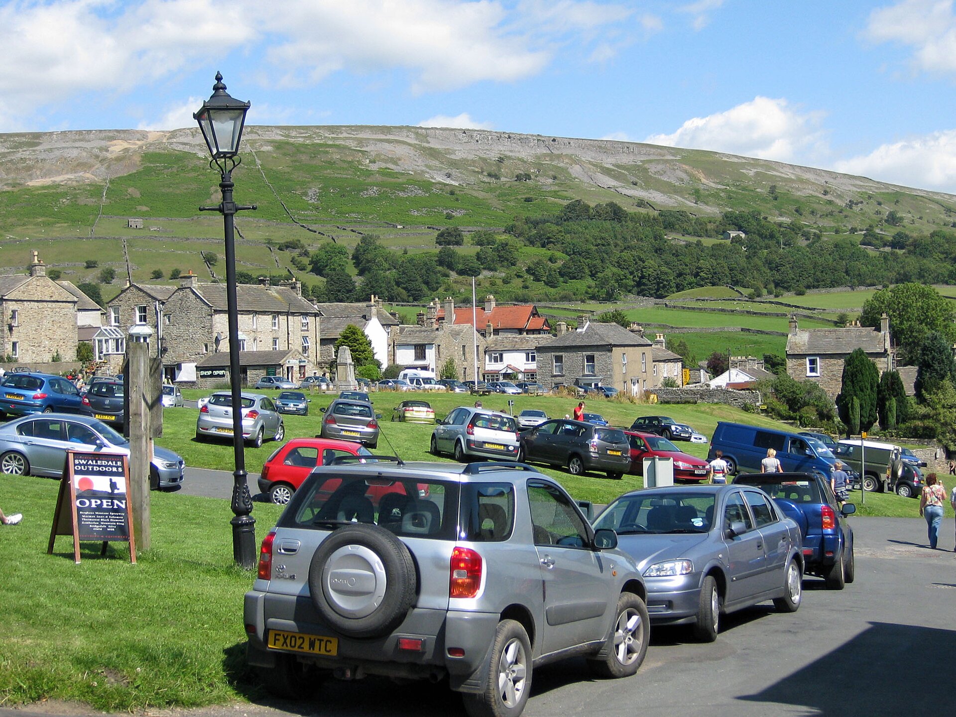Na zdjęciu widoczny jest teren wiejski. Na pierwszym planie znajduje się dużo zaparkowanych wzdłuż ulicy i na trawnikach samochodów. W tle widoczne są zabudowania i  kamienne domy ze spadzistymi dachami. Dalej widoczny jest teren górski. Łagodny stok pokryty jest drzewami i trawą. Nad stokiem widać niebieskie niebo z białymi chmurami
