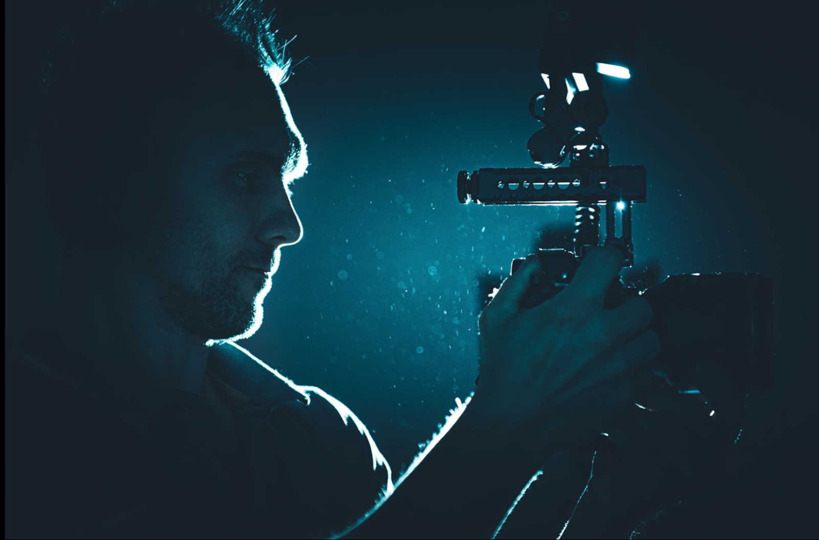 Zdjęcie przedstawia mężczyznę w ciemnym pomieszczeniu z aparatem fotograficznym w rękach. Kadr obejmuje głowę z profilu, ramiona i ręce z aparatem uniesione na wysokość twarzy. Mężczyzna patrzy na aparat, z pewnej odległości, lekkie oświetlenie rysuje jedynie kontur jego twarzy, rąk i aparatu fotograficznego.