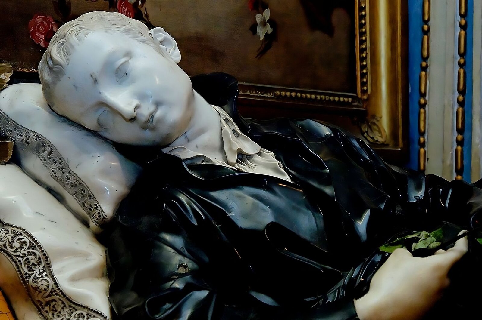 Zdjęcie przedstawia rzeźbę leżącego mężczyzny. Jest młody, ubrany w koszulę luźno rozpiętą pod szyją i sutannę z wysokim kołnierzem. Jego głowa spoczywa na poduszkach, przechylona na prawy bok. Prawa ręka spoczywa na brzuchu. W tle widać zdobioną ramę obrazu i ozdobną sztukaterię na ścianie.