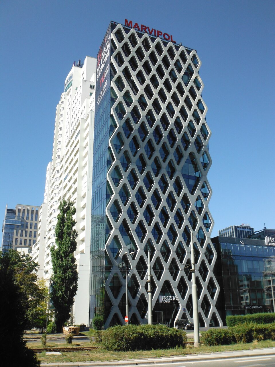 Zdjęcie przedstawia Prosta Tower w Warszawie. Wieżowiec wykonany jest ze szkła. Na jednej, całej ścianie umieszczona jest ażurowa, w kształcie rombów, fasada z żelbetonu.