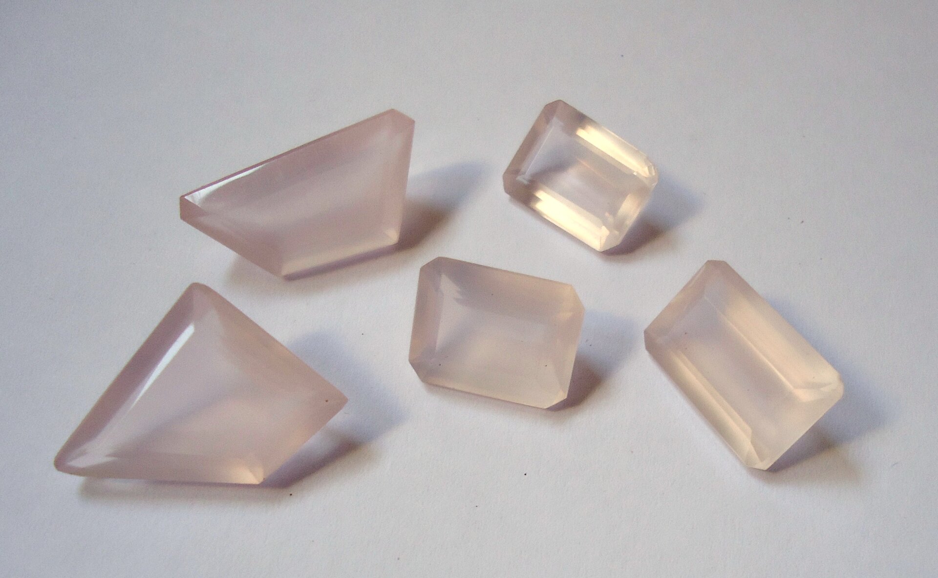 Zdjęcie przedstawia pięć oszlifowanych kawałków kwarcu różowego o mlecznoróżowej barwie. Trzy mają szlif szmaragdowy, czyli prostokątny, zaś dwa są nieregularnymi pięciobokami. Wszystkie kamienie leżą obok siebie na białym tle.
