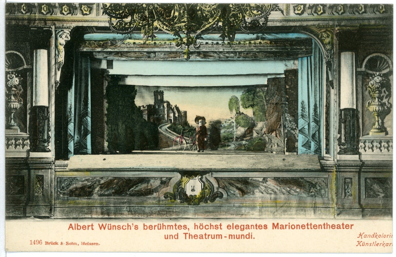 Pocztówka przedstawia teatr marionetek. Jest ukazana scena, na której znajduje się jedna marionetka stojąca na tle krajobrazu i dużego zamku. Pod pocztówką widnieje napis w języku niemieckim.