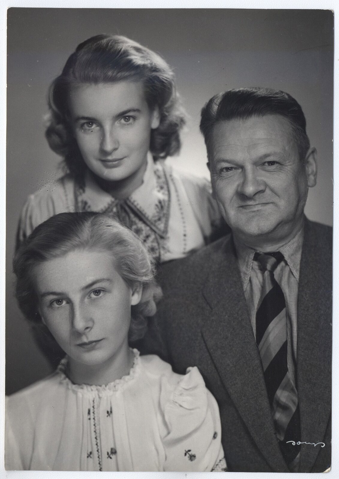 Czarno‑białe zdjęcie portretowe trojga ludzi. Po lewej dwie młode kobiety o jasnych włosach sięgających ramion. Kobieta znajdująca się poniżej ma na sobie białą bluzkę ozdobioną haftowanymi kwiatkami, z bufiastymi rękawkami, wiązaną pod szyją na tasiemkę. Pochyla lekko głowę w lewą stronę, ma poważny wyraz twarzy. Kobieta znajdująca się powyżej ma na sobie zdobioną kwiatowym haftem bluzkę z kołnierzykiem. Lekko uśmiecha się. Po prawej znajduje się uśmiechnięty mężczyzna w średnim wieku. Jego włosy są ciemne, krótkie i zaczesane do góry. Ma na sobie koszulę z kołnierzykiem, krawat w ciemne, skośne pasy i ciemną marynarkę.