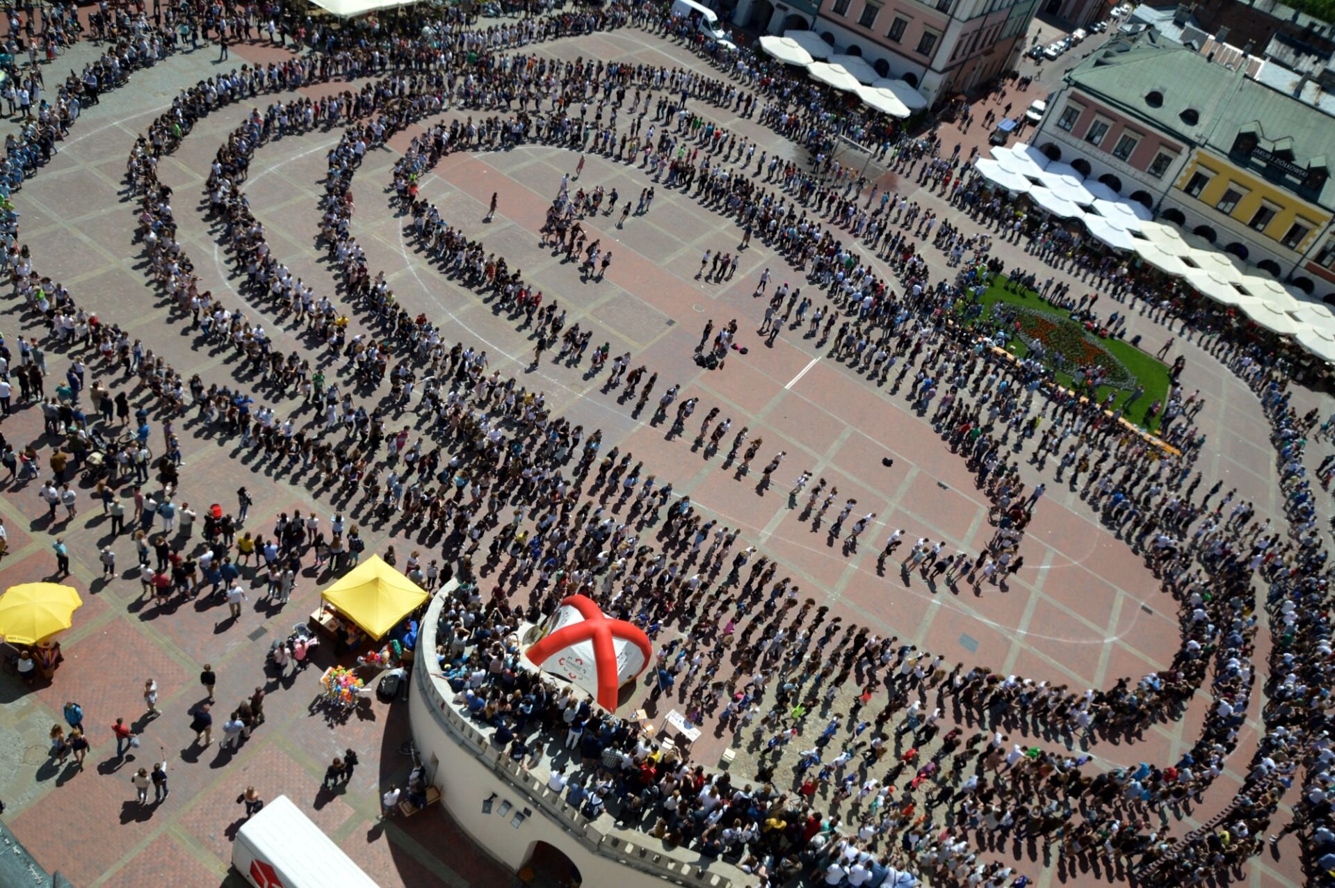 Fotografia prezentuje bicie rekordu w liczbie uczestników biorących udział w tańcu zwanym „Belgijką” na Rynku Wielkim w Zamościu. Fotografia została wykonana z lotu ptaka. Na rynku widoczna jest bardzo duża grupa osób, które stoją parami, tworząc kształty przypominające spłaszczone elipsy. Jest ich pięć – w środku najmniejsza oraz kolejne cztery, coraz większe. Na zewnątrz obecni się obserwujący. W tle fotografii uchwycono miejskie budynki oraz ustawione przed nimi chroniące przed słońcem parasole.  