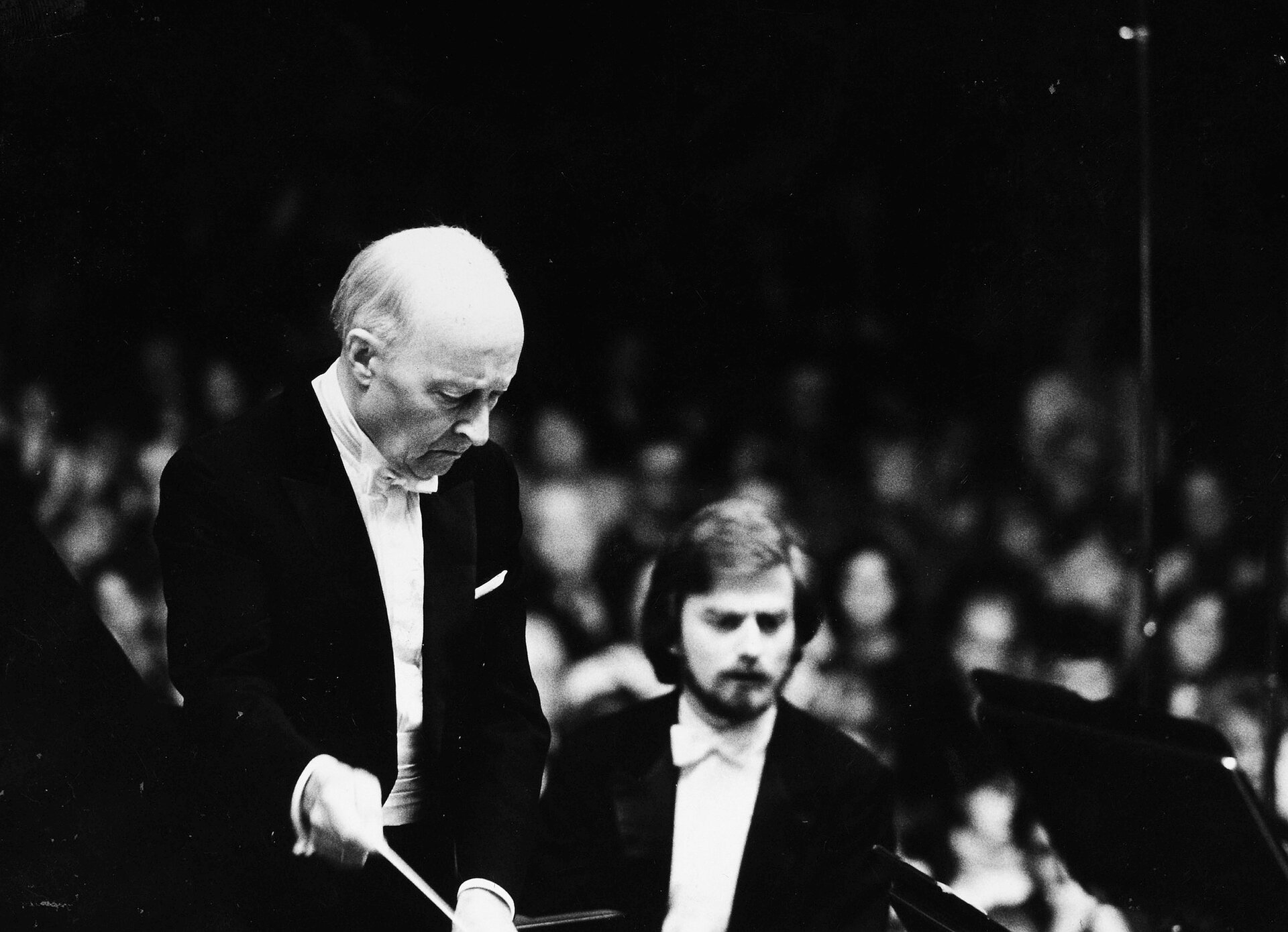 Na czarno-białym zdjęciu znajduje się Witold Lutosławski ukazany jako dyrygent. Ubrany jest w białą koszulę, czarny frak, pod szyją ma białą muchę, Kompozytor w ręku trzyma batutę. W tle widać niewyraźne sylwetki wielu ludzi.