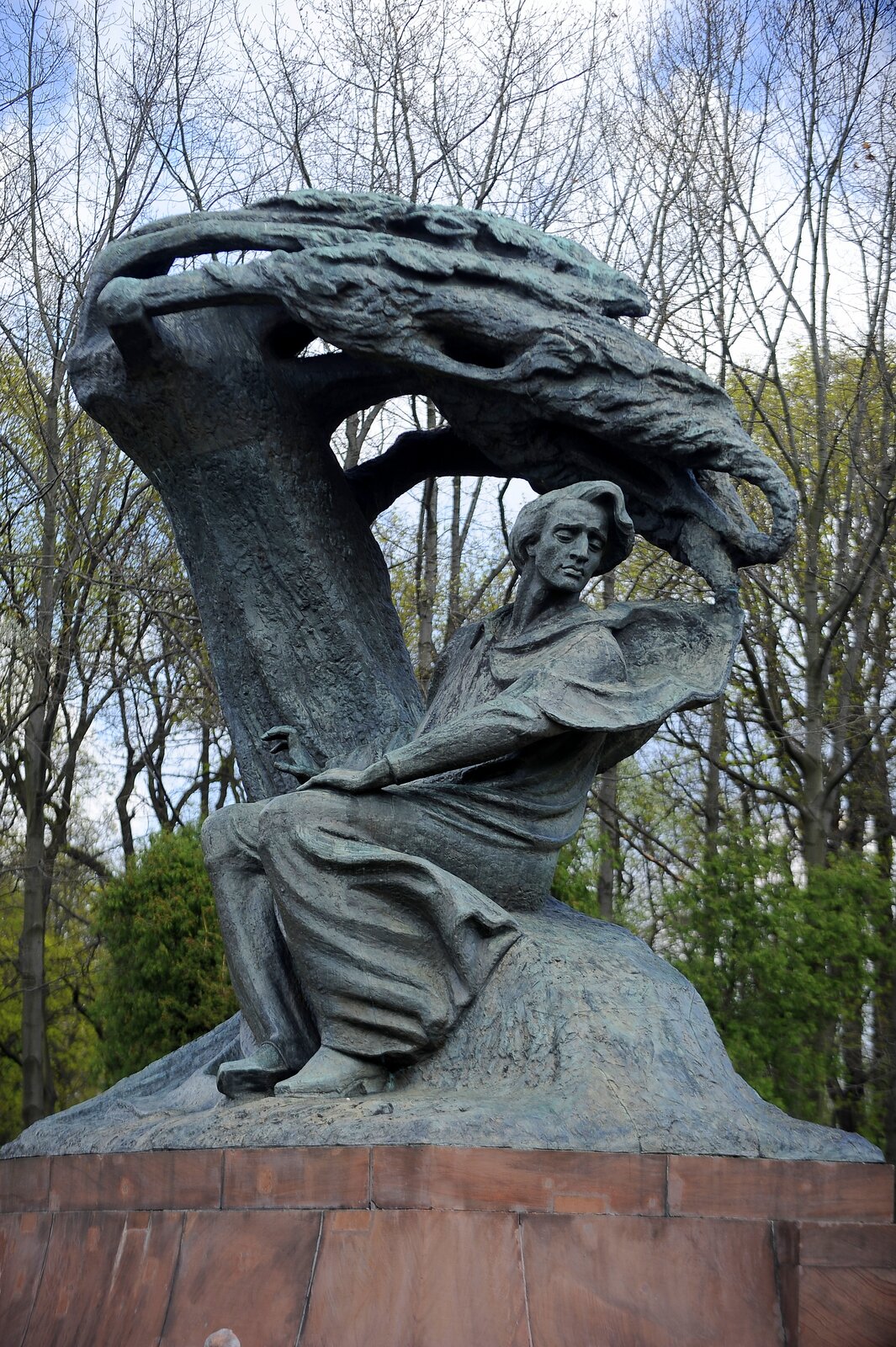 zdjęcie przedstawia pomnik siedzącego mężczyzny, palce jego lewej reki sa rozpostrarte jak do gry na fortepianie, nad nim pochyla się płacząca wierzba