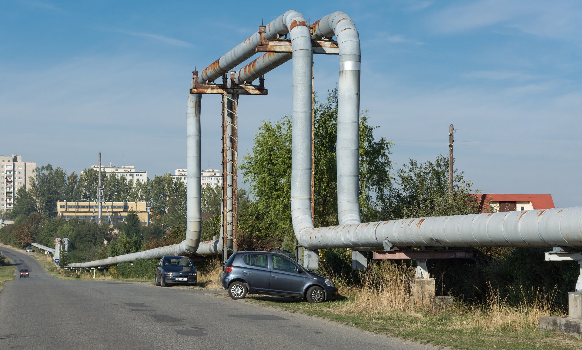 Rys. 5. Na zdjęciu przedstawiony jest fragment rurociągu ciepłowniczego. Rurociąg to rura w kształcie cylindra. Średnica tej rury jest porównywalna z długością od ramienia do końca dłoni. Rura wykonana jest z metalu w szarym kolorze. Przez pewien odcinek rura biegnie poziomo, równolegle do ziemi i jest umieszczona na powtarzających się co jakiś czas betonowych wspornikach zakończonych poziomymi, metalowymi ramionami. W pewnym momencie rura zakrzywia się i zaczyna biec pionowo w górę. Następnie, na pewnej wysokości nad ziemią rura znowu zakrzywia się i zaczyna biec poziomo. Na tym odcinku rurę podtrzymują dwa metalowe słupy zakończone poziomymi ramionami. Po przebyciu pewnej odległości rura zagina się pionowo w dół i opada do tej samej wysokości, którą miała na początkowym odcinku. Dalej rura biegnie znowu poziomo i ponownie jest podparta na betonowych wspornikach zakończonych poziomymi, metalowymi ramionami. Rura znajduje się przy miejskiej ulicy, przy niej zaparkowane są dwa samochody.