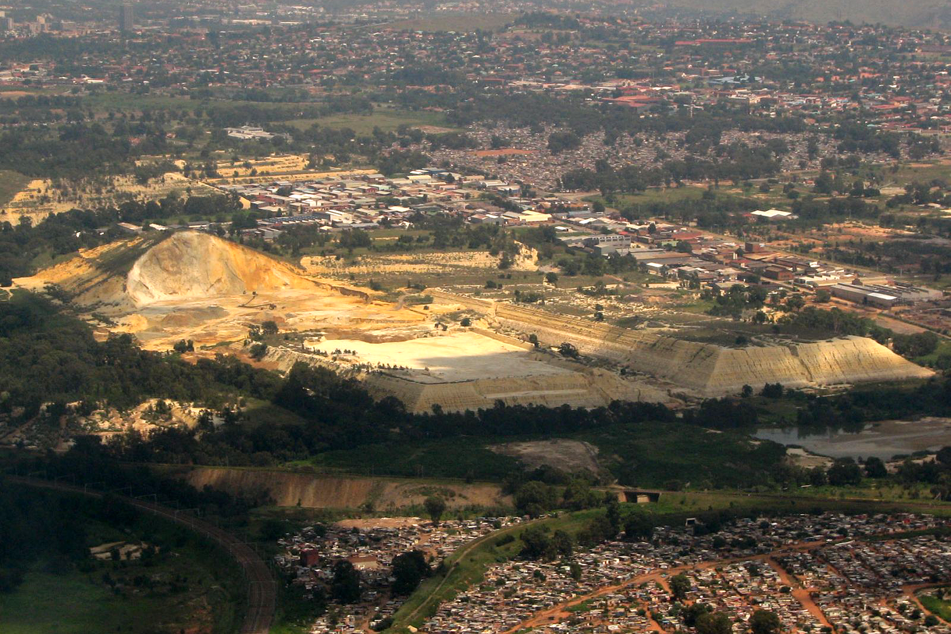 Fotografia przedstawia pejzaż przemysłowy, w ujęciu z góry. W centrum znajdują się żółte i kremowe bryły; to hałdy składowanych odpadów. Są ogromne w porównaniu z domami w okolicy.