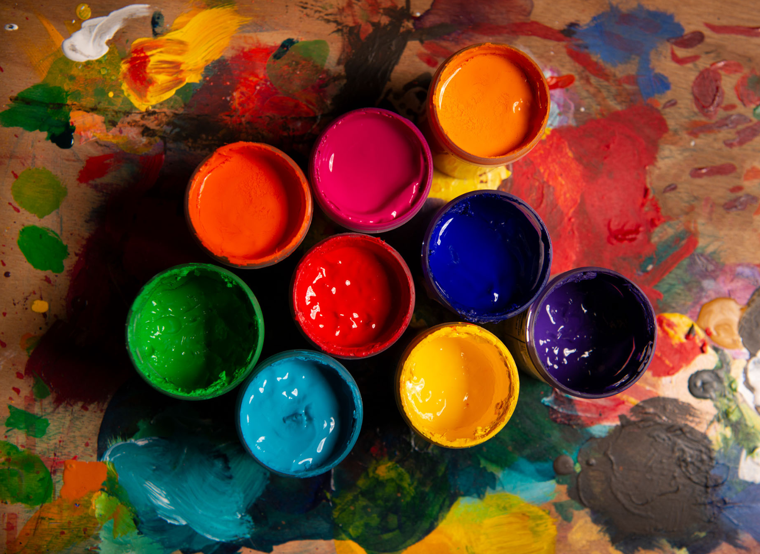 Zdjęcie wykonane jest z góry i przedstawia farby plakatowe w otwartych słoiczkach. Są gęste, mają  barwy ciepłe i zimne. Stoją na papierze pomalowanym w plamy, jest ich 9.