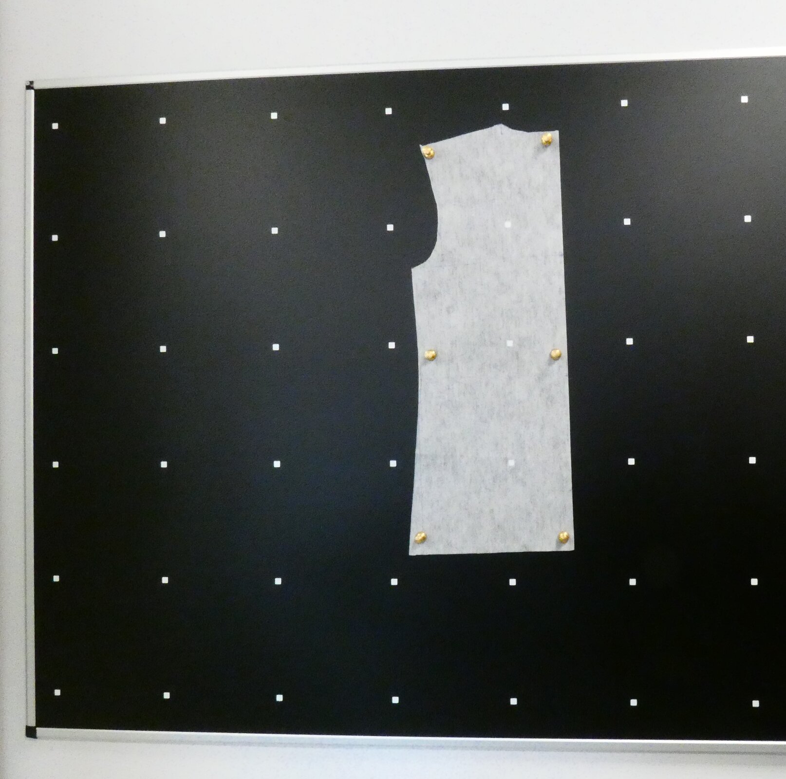 Zdjęcie przedstawia dużą czarną tablicę, na której symetrycznie rozmieszczone są białe punkty. W jej centralnej części rozwieszony jest kawałek białego materiału. Jest przyczepiony do tablicy za pomocą pinezek.