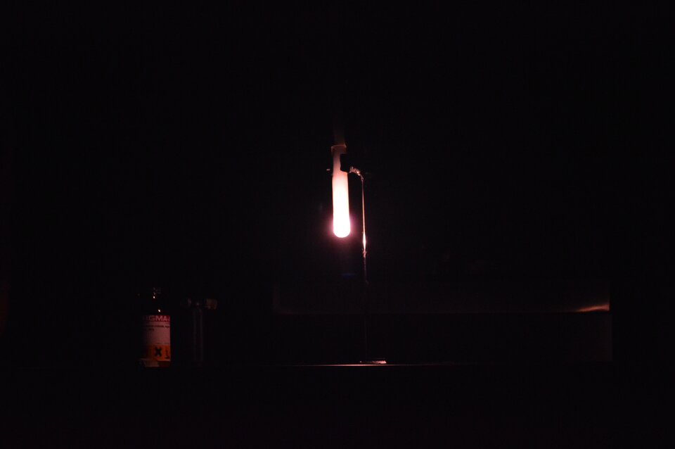 Zdjęcie przedstawia ciemne pomieszczenie, w którym znajduje się świecący płomień zamknięty w szklanym naczyniu. Płomień jest wynikiem reakcji spalania mieszaniny chloranu(pięć) potasu i cukru.