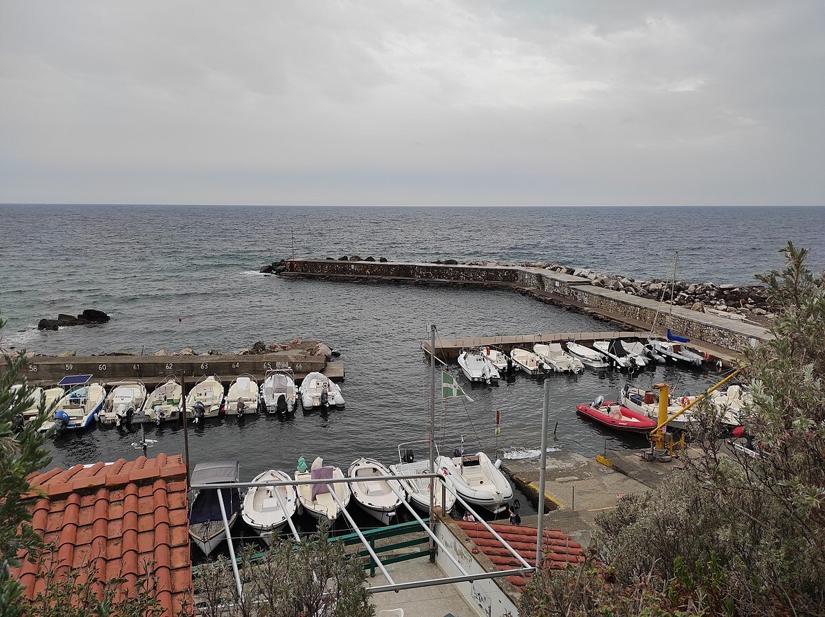 Kliknij aby powiększyć Zdjęcie przedstawia marinę, czyli postój dla łodzi. Na wprost widoczny falochron na dużym akwenie. Niebo zachmurzone, szare.