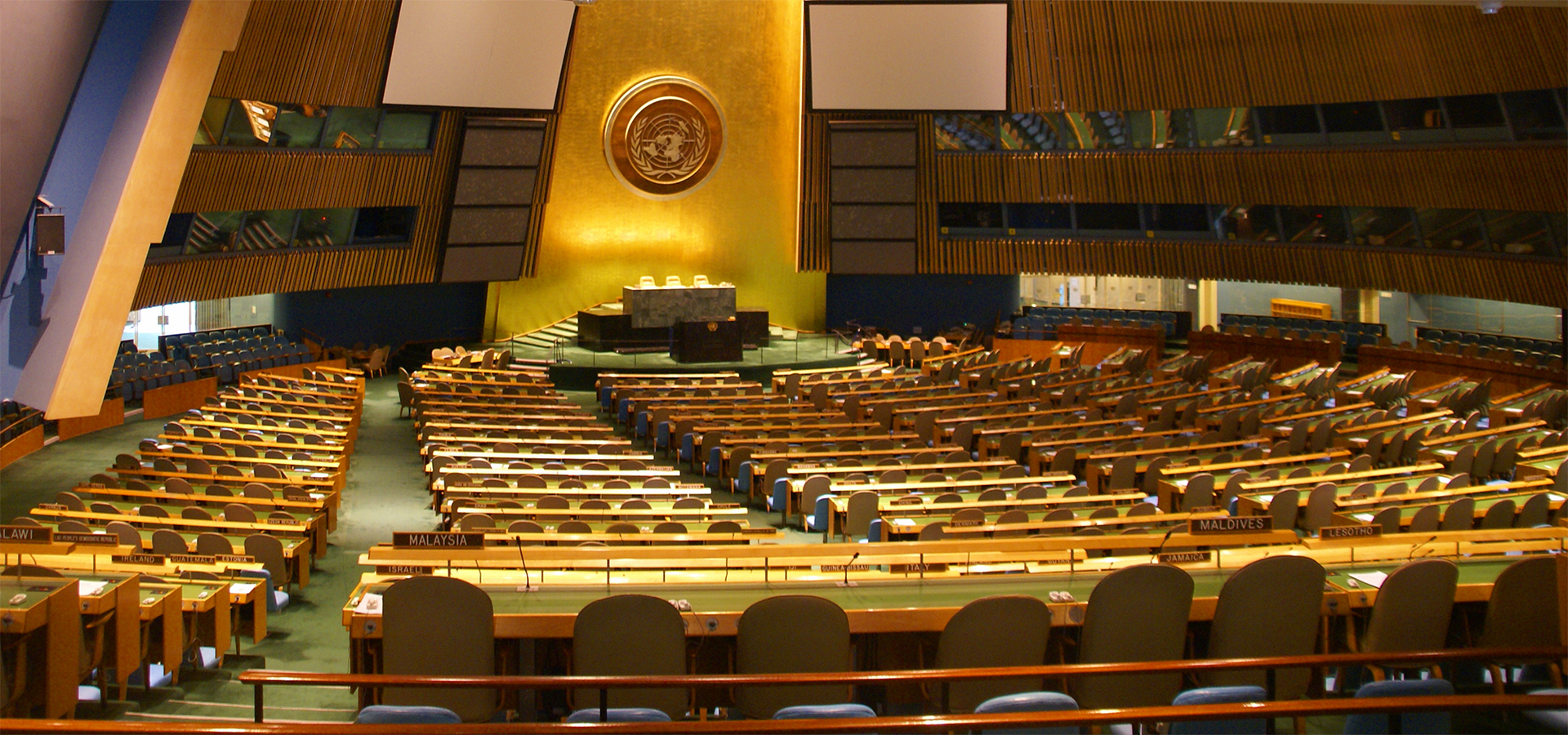 Zdjęcie przedstawia pomieszczenie o kształcie owalnym. Widać na nim drewniane siedzenia ustawione w rzędach. W tle widać podest i ustawiony na nim stół oraz trzy krzesła.