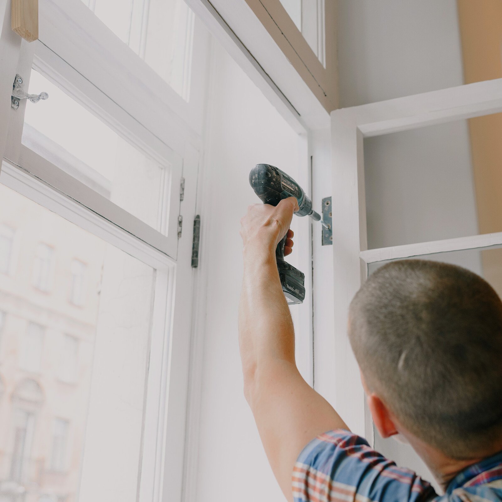 Na zdjęciu widoczny jest mężczyzna montujący w mieszkaniu zawiasy do okna. 