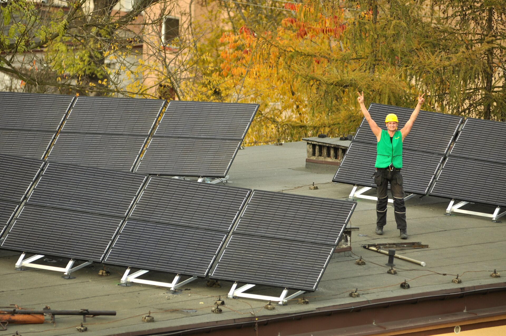 Zdjęcie przedstawia panele fotowoltaiczne umieszczona w trzech modułach na dachu. Między nimi stoi mężczyzna w czarnych spodniach, zielonej kamizelce i żółtym kasku na głowie z uniesionymi rękami do góry.