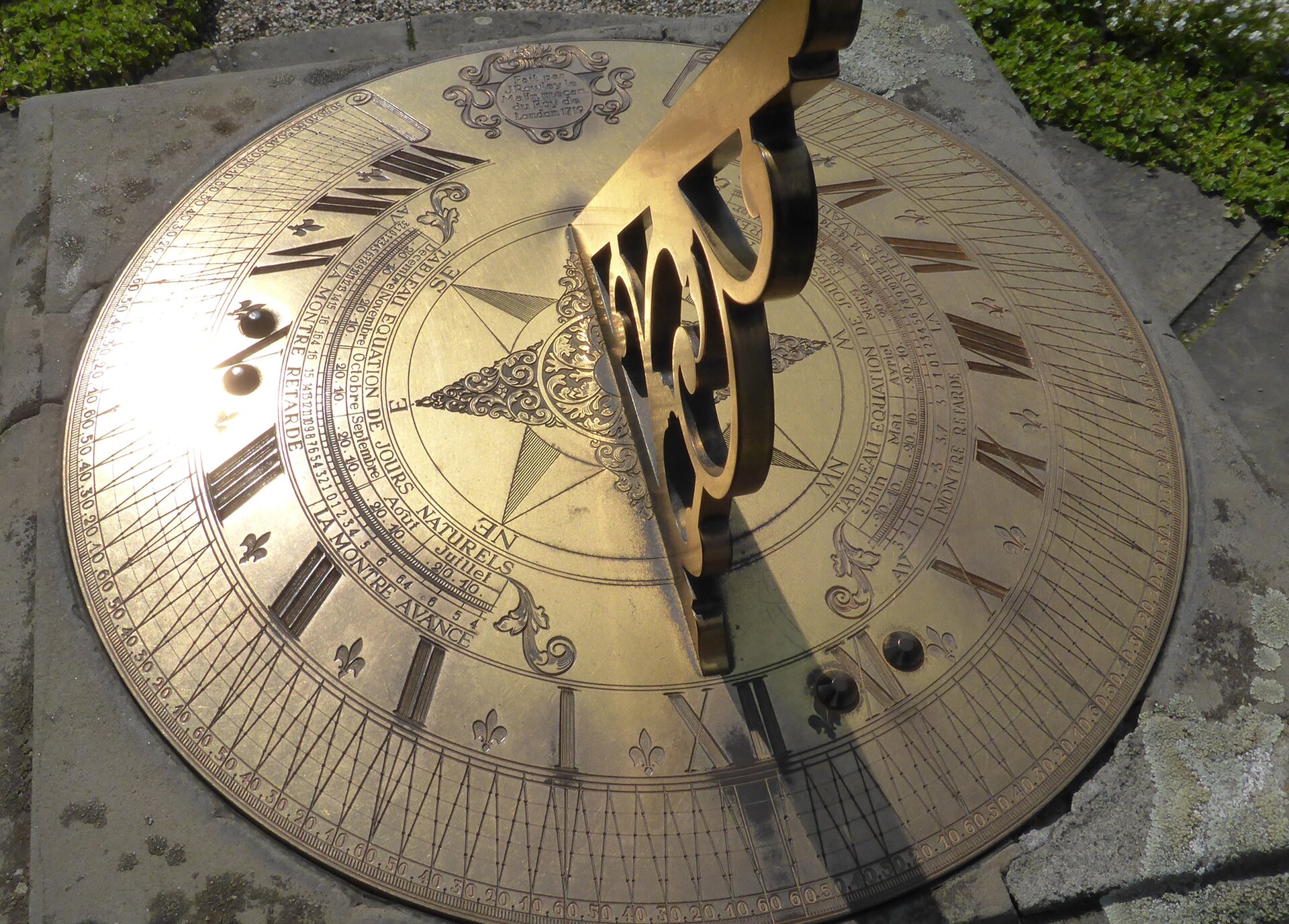 Na zdjęciu lekko wypukła okrągła tarcza zegara słonecznego sfotografowana z góry. Umieszczona jest na ziemi na kamiennych płytach, w tle widać fragment trawnika. Zegar wykonany jest z metalu w lekko złotym kolorze, z lewej strony widać, jak błyszczy się od słońca. Na środku umieszczony jest gruby pręt (gnomon) z ozdobną końcówką, przestrzeń kąta nachylenia między nim a płaszczyzną tarczy wypełnia fantazyjna ozdoba, również wykonana z metalu. Przypomina rzeźbione drewniane ozdoby na zabytkowych budynkach. Na środku tarczy umieszczona jest ośmioramienna gwiazda wskazująca główne i pośrednie kierunki świata. Zdjęcie wykonano z drugiej strony zegara, dlatego na pierwszym planie widać rzymską liczbę XII do góry nogami, po czym I i kolejne liczby  są po lewej stronie,  w miejscu po liczbie VIII jest przerwa na tarczy. Umieszczono tam małą okrągłą tabliczkę ozdobioną koroną i motywami roślinnymi, jest tam m.in. napis "London, 1719". Za tabliczką ciąg liczb jest kontynuowany aż do XII. Na zewnętrznej stronie tarczy między kolejnymi liczbami są wygrawerowane arabskie liczby od 10 do 60. Wszystkie ozdoby i napisy  (w języku francuskim) na tarczy są artystycznie i starannie wygrawerowane. 