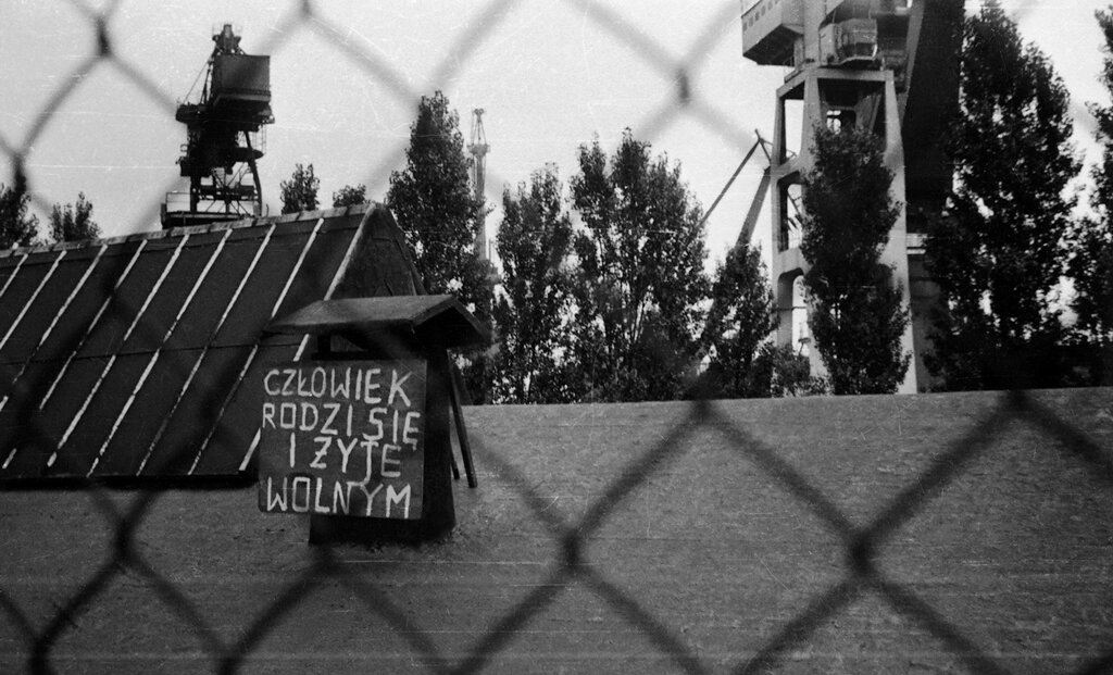 Czarno-biała fotografia dachów budynków stoczni w Gdańsku wykonana przez płot z siatki. Znajduje się na niej transparent "człowiek rodzi się i żyje wolnym".