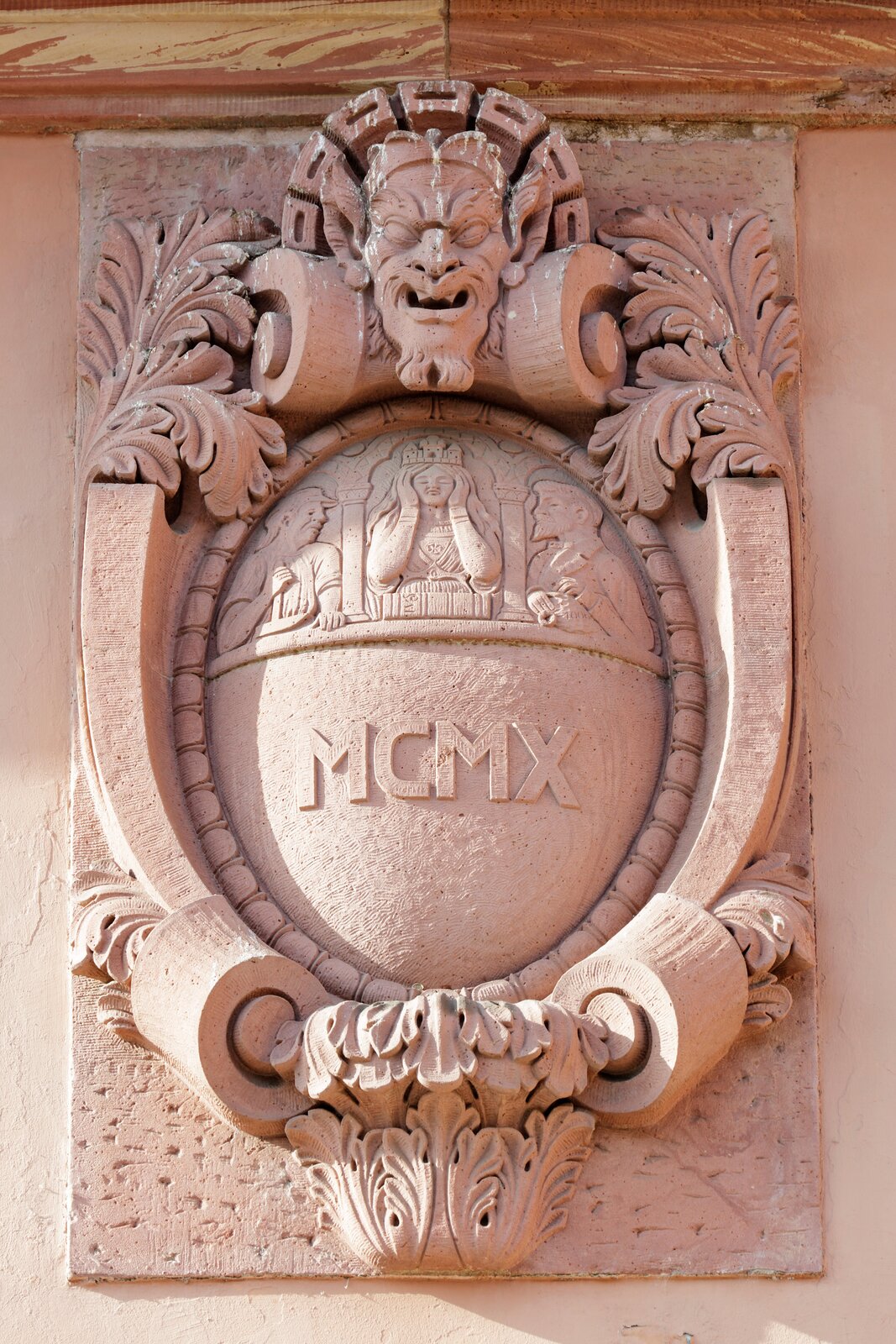 Ilustracja przedstawia ornament – kartusz. Element jest przymocowany do ściany i przedstawia głowę człowieka z otwartymi ustami. Poniżej głowy znajduje się wypukły element z płaskorzeźbą oraz z napisem MCMX.