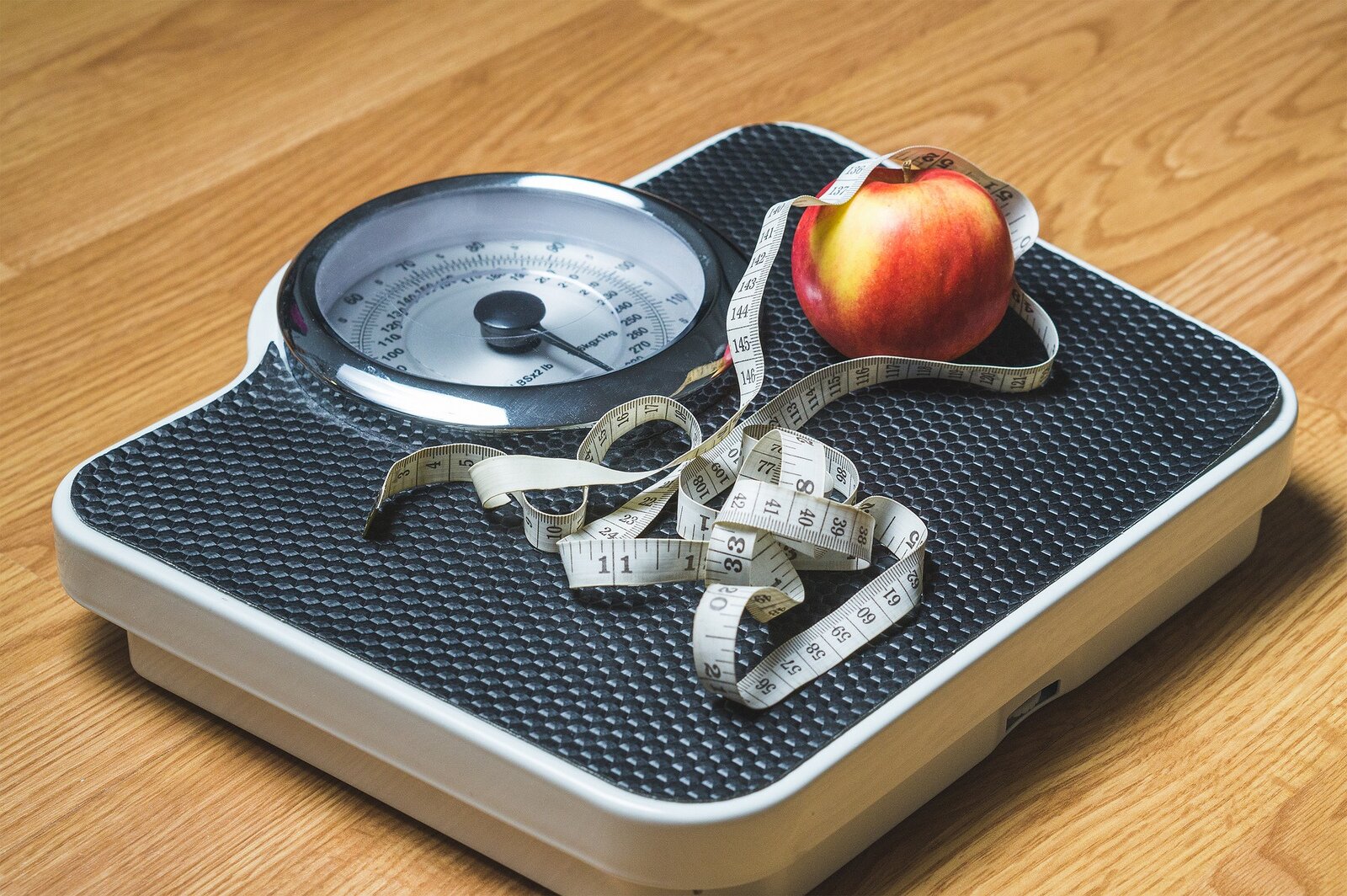 Zdjęcie przedstawia wagę, na której leży zwinięty centymetr oraz jabłko.
