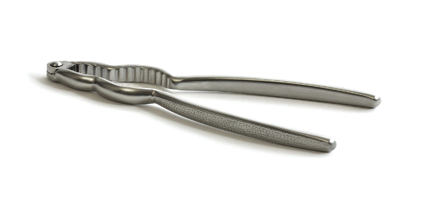 Fotografia przedstawia położonego na białym blacie metalowe narzędzie ręczne kuchenne. Jest to dziadek do orzechów. Narzędzie składa się z dwóch ramion z cienkim prostym uchwytem połączonych zaokrąglonymi końcówkami.  Wewnątrz para ramion ma dwa łuki: u góry mniejsze, niżej większe z poprzecznymi wyżłobieniami .