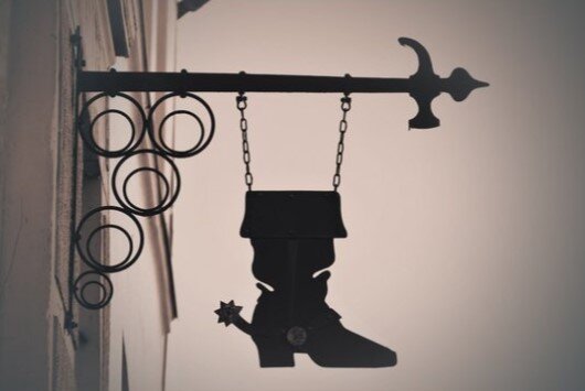 Fotografia przedstawia fragment kamienicy z szyldem warsztatu rzemieślniczego szewca. Do ściany kamienicy przytwierdzono poziomo pręt w kształcie ozdobnego młotka. Od trzonka młotka zwisa metalowy, płaski but z cholewą, obcasem i ostrogą w kształcie gwiazdki. Za butem, przy ścianie znajdują się ozdobne obręcze różnej wielkości.