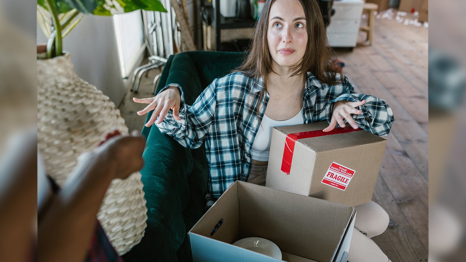 Zdjęcie przedstawia młodą, ciemnowłosą kobietę, która siedzi na sofie, trzymając na kolanach pudełko zaklejone czerwoną taśmą, na boku którego jest naklejka z napisem "PLEASE HANDLE WITH CARE. FRAGILE." Na siedzisku obok brunetki leży drugie pudełko. W lewym dolnym rogu kadru jest ukazana prawa ręka osoby stojącej przodem do kobiety na sofie, której wzrok jest skierowany w prawą stronę w górę. 